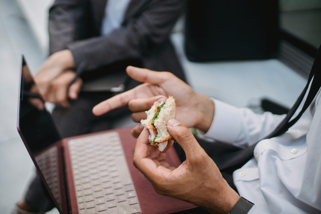 Close-up of a professional holding a sandwich during a work break with a laptop nearby.