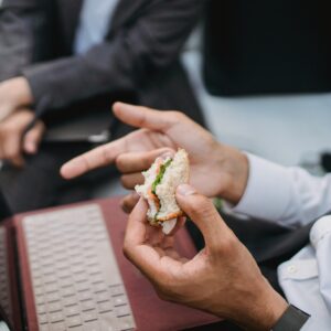 Close-up of a professional holding a sandwich during a work break with a laptop nearby.